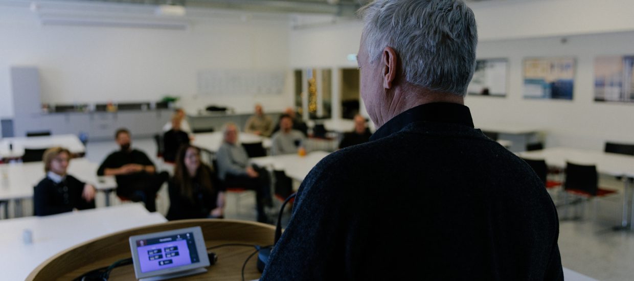 Mann med kort grått hår sett bakfra. Snakker til forsamling i et konferanserom.